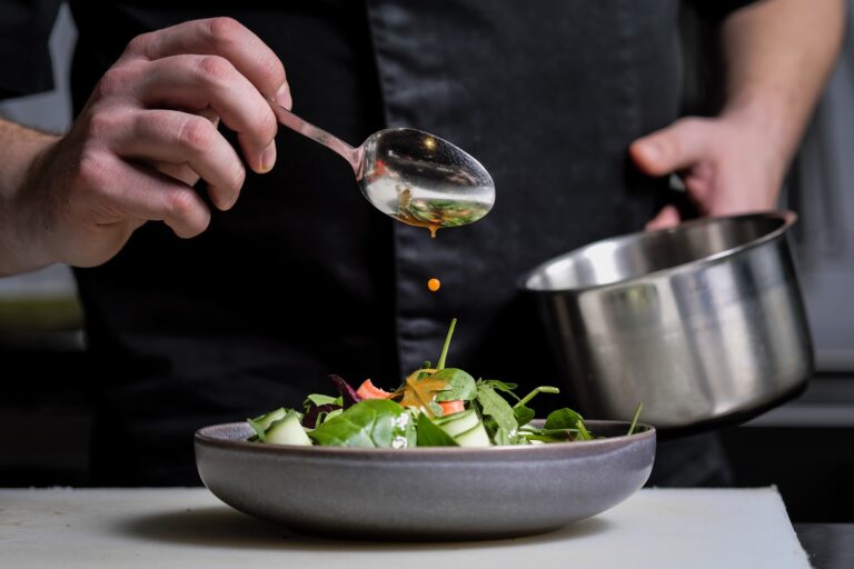 Close up of the hands of a male chef on a black background. Pour sauce from the spoon on the salad dish.