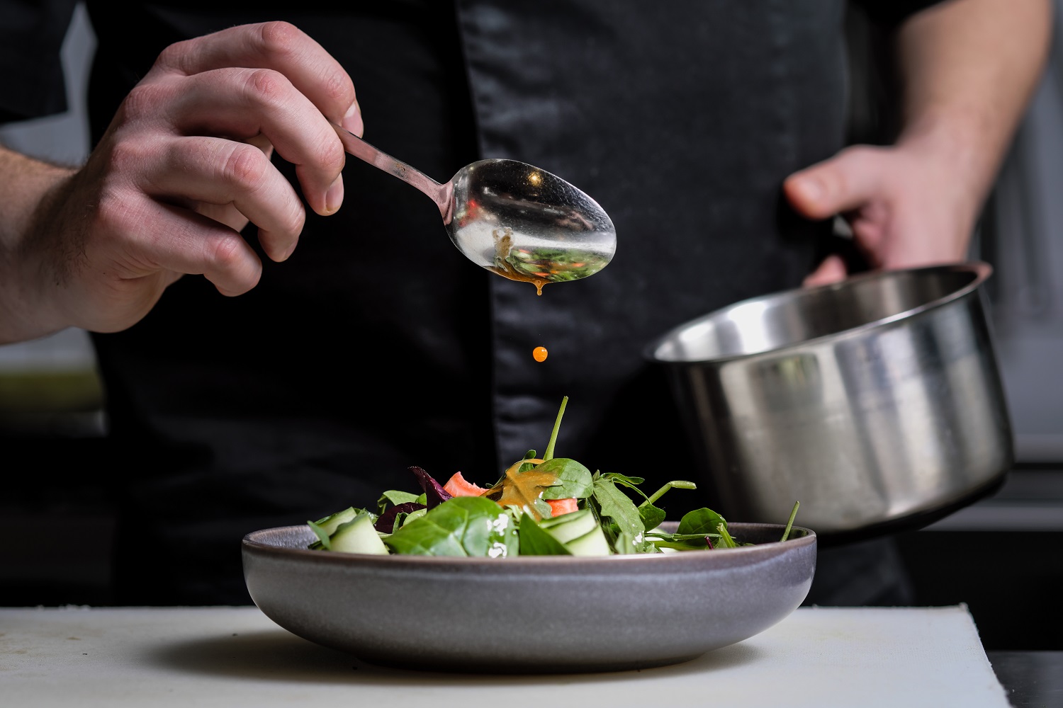 Close up of the hands of a male chef on a black background. Pour sauce from the spoon on the salad dish.